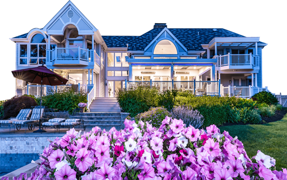 The back of a large two-story beach house lit up from inside with a large LifeRoom outdoor living room attached to it with the screens open with a pool, bushes, and purple flowers in the foreground.